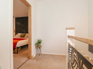 A hallway with beige carpet and a small plant on a stand next to an open door showing a bed with a red and orange blanket at Yamthwaite in Grizedale near Hawkshead