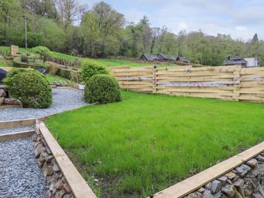 A garden with grass and gravel pathways bordered by wooden edges and bushes with a wooden fence and trees in the background at Yamthwaite Grizedale near Hawkshead