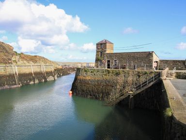 A stone building on a harbor with calm water and rocky cliffs at The Hawthorns in Llanfechell