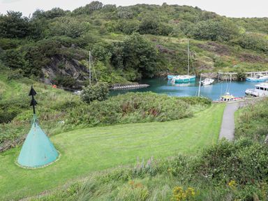 A grassy area leading to a small harbor with boats and surrounding hills at The Hawthorns in Llanfechell