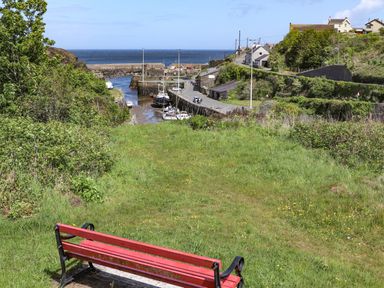 A grassy hill with a red bench overlooking a small harbor with boats and houses near the sea at The Hawthorns in Llanfechell