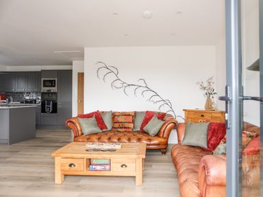 A living room with two brown leather sofas and a wooden coffee table next to a kitchen with grey cabinets at The Hawthorns in Llanfechell
