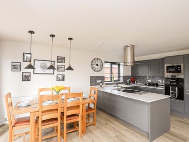 A kitchen and dining area with a wooden dining table and six chairs next to a gray kitchen island with modern appliances at The Hawthorns in Llanfechell
