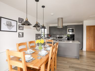 A dining area with wooden table set for six and kitchen with grey cabinets and island at The Hawthorns in Llanfechell