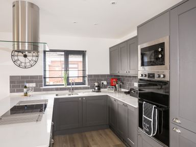 A kitchen with gray cabinets a double sink an oven and microwave a stovetop with a stainless steel hood and a window with blinds at The Hawthorns in Llanfechell