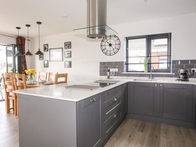 A kitchen with gray cabinets a white countertop a dining table with wooden chairs and yellow flowers at The Hawthorns in Llanfechell