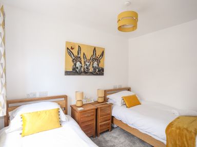 A bedroom with two single beds wooden bedside cabinets yellow lamps and a painting of three donkeys at The Hawthorns in Llanfechell
