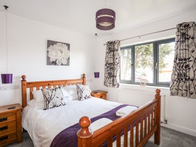 A bedroom with a wooden bed frame white bedding purple accents and floral curtains at The Hawthorns in Llanfechell