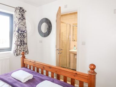 A bedroom with a wooden bed, purple blanket, white towels, a window with floral curtains, a round wall mirror, and an open door leading to a bathroom at The Hawthorns in Llanfechell