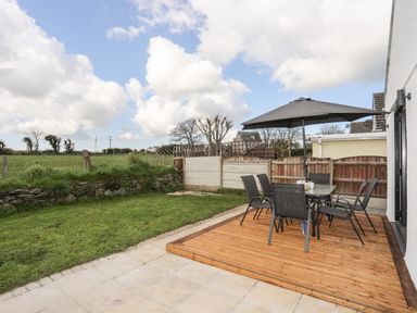 A patio with a table and six chairs under a large umbrella next to a grassy lawn at The Hawthorns in Llanfechell