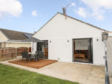 A patio with outdoor dining table and umbrella next to a lawn and white house with open doors at The Hawthorns in Llanfechell