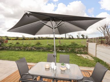 An outdoor patio with a glass table and four chairs under a large umbrella overlooking a grassy field at The Hawthorns in Llanfechell