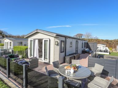 An outdoor patio area with a table and chairs a barbecue and seating in front of a beige modular home at 97 Crugan in Llanbedrog