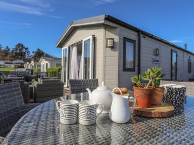An outdoor patio with a glass table and wicker chairs in front of a beige caravan at 97 Crugan in Llanbedrog