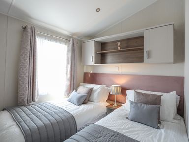 A bedroom with two single beds side by side a table lamp between them a window with curtains and overhead cabinets at 97 Crugan in Llanbedrog