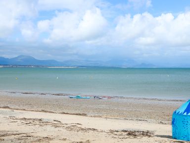 A sandy beach with scattered seaweed a blue tent and water sports equipment near the shore with mountains and clouds in the background at 97 Crugan in Llanbedrog