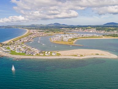 An aerial view of a coastal town with houses, a marina with boats, a sandy beach, and mountains in the background at 97 Crugan in Llanbedrog