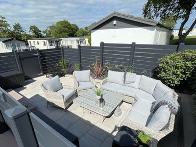 An outdoor patio area with grey cushioned wicker furniture a glass coffee table plants and a dark grey fence at 97 Crugan in Llanbedrog