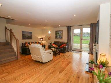 A living room with brown sofas a white armchair wooden floor and glass doors leading outside at Barn End 9 Stad Clynnog in Newborough