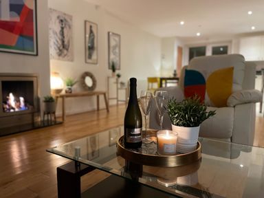 A living room with a bottle, glasses, candle, and plant on a glass table near an armchair with colorful pillows and a fireplace at Barn End, 9 Stad Clynnog in Newborough