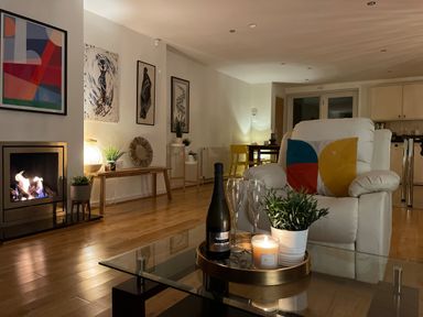 A living room with a lit fireplace a white armchair and a glass coffee table with a candle plant and bottle at Barn End 9 Stad Clynnog in Newborough