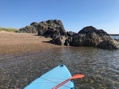 The front of a blue kayak with a red paddle on clear water near a rocky shoreline with a sandy beach and grass under a clear sky at Barn End 9 Stad Clynnog in Newborough
