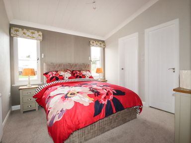 A bedroom with a double bed with floral red bedding between two windows and two white doors at Acorn Lodge in St Day