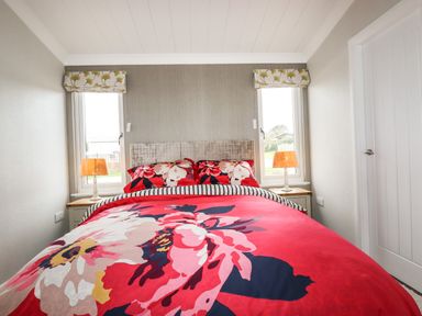 A bedroom with a bed with floral bedding two bedside tables with orange lamps and two windows at Acorn Lodge in St Day