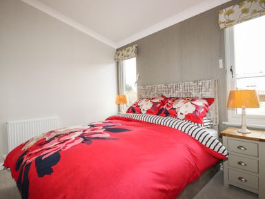 A bedroom with a bed with red floral bedding two bedside tables with lamps and two windows at Acorn Lodge in St Day