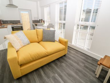 A yellow sofa with pillows in a room with large windows and a kitchen and dining area visible at Perran Pearl A in Perranporth