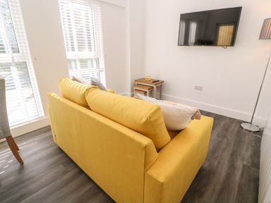 A yellow sofa with cushions in a living room with a wall-mounted TV and a small wooden nesting table at Perran Pearl A in Perranporth