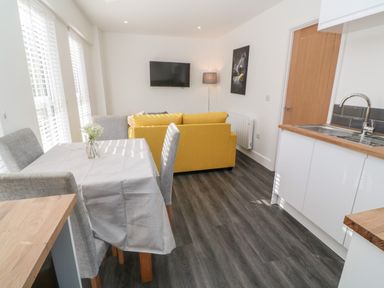 A dining table with four chairs next to windows and a yellow sofa facing a wall-mounted TV in a living room with a kitchen sink in Perran Pearl A in Perranporth