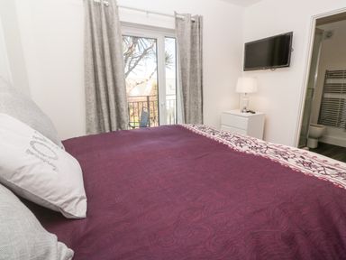 A bedroom with a bed covered in a purple and white bedspread a bedside table with a lamp a wall-mounted TV and an open door to a bathroom at Perran Pearl A in Perranporth