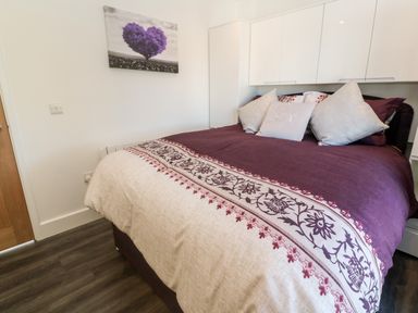 A bedroom with a bed covered in purple and beige bedding and decorated with four pillows and a wall painting of a purple heart-shaped tree at Perran Pearl A in Perranporth