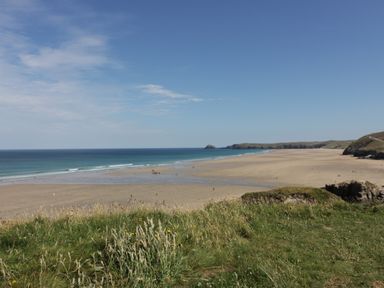 A wide sandy beach with ocean waves and grassy cliffs in the foreground at Perran Pearl B in Perranporth