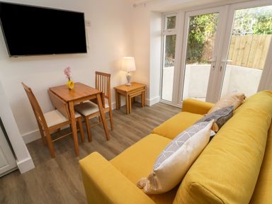A room with a yellow sofa with pillows a small wooden dining table with two chairs a nested side table with a lamp and glass doors leading outside at Perran Pearl B in Perranporth