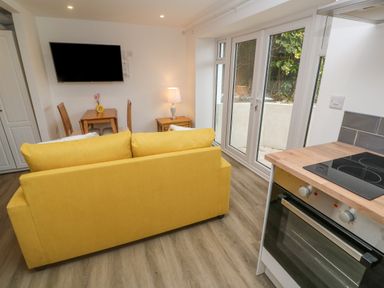 A living area with a yellow sofa, dining table, wall-mounted TV, lamp, and kitchen oven with glass doors at Perran Pearl B in Perranporth