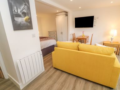 A living room with a yellow sofa, wall-mounted TV, wooden table and chairs, lamp, and view into bedroom with bed and wardrobe at Perran Pearl B in Perranporth