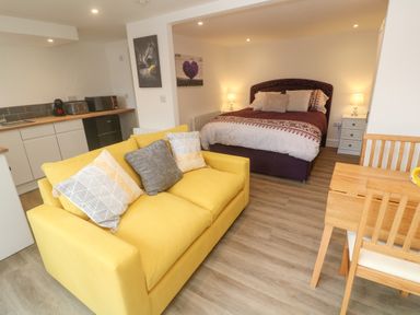 An open plan room with a yellow sofa a bed with bedside tables and lamps a kitchenette and a wooden dining table with chairs at Perran Pearl B in Perranporth