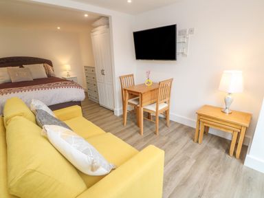 A room with a yellow sofa a bed a wooden dining table with two chairs a wall-mounted TV and a side table with a lamp at Perran Pearl B in Perranporth