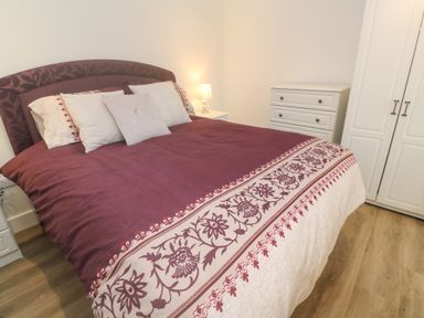 A bedroom with a bed covered in a patterned duvet several pillows a bedside lamp a white dresser and a white wardrobe at Perran Pearl B in Perranporth