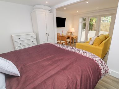 A bedroom with a bed a white wardrobe a chest of drawers a wooden table with chairs a yellow sofa and glass doors at Perran Pearl B in Perranporth