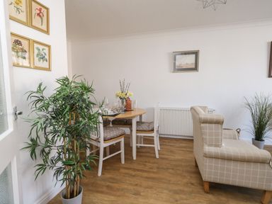 A dining area with a round table set with glassware and white chairs beside a beige checkered sofa in a room with wooden flooring and wall art at Beach Side in West Bay