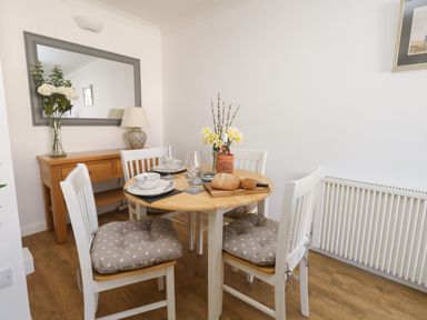 A dining area with a round wooden table set with bowls glasses and bread surrounded by four white chairs with cushions and a wooden sideboard with a lamp and flowers at Beach Side in West Bay