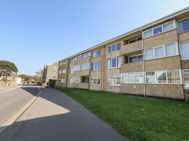 A street view of a three-story residential building with windows and balconies next to a sidewalk and a grass lawn at Beach Side in West Bay