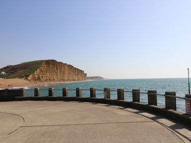 A concrete promenade with a railing overlooking the sea and cliffs with grassy tops at Beach Side in West Bay