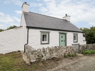 A white stone cottage with two chimneys and green door surrounded by a low stone wall at Rhytty Bach in Ty Croes near Rhosneigr