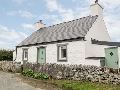 A white stone cottage with green doors and windows behind a low stone wall at Rhytty Bach in Ty Croes near Rhosneigr