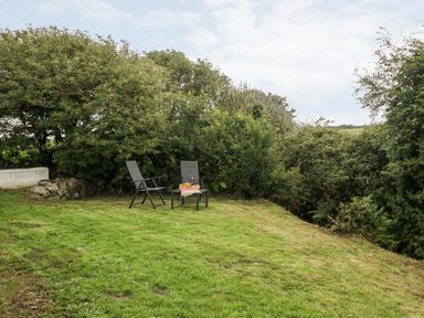 A grassy garden area with two black chairs and a small table with items near dense bushes at Rhytty Bach in Ty Croes near Rhosneigr