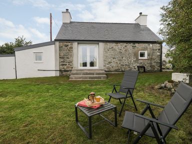 A stone cottage with a lawn and two chairs with a table in the garden at Rhytty Bach in Ty Croes near Rhosneigr